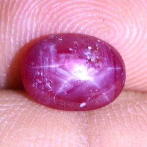 Può includere: Primo piano di una gemma di rubino ovale con taglio cabochon. Il rubino rosso intenso mostra un effetto stella a sei raggi, noto anche come asterismo. La superficie presenta piccole inclusioni e una finitura lucida.
