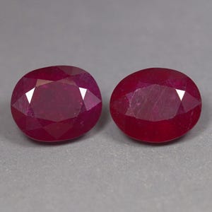 May include: Two oval-shaped, faceted rubies with a deep, saturated red colour. Each gemstone features a polished surface with multiple triangular and trapezoidal facets that reflect light, showcasing a translucent quality and a vibrant crimson hue.