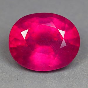May include: A vibrant, oval-cut gemstone with a deep, saturated magenta-pink hue. The stone features a faceted surface that reflects light, creating a brilliant, sparkling appearance across its polished, symmetrical, and rounded oval shape.