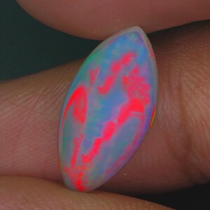 May include: A polished, oval-shaped opal gemstone. The opal displays a vibrant play-of-color, with flashes of red, blue, and green against a milky white base. The gemstone is held between fingers, showcasing its size and shape.