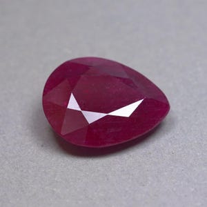 Puede incluir: Una piedra preciosa facetada en forma de pera con un color rojo magenta profundo y saturado. La piedra presenta una superficie pulida con múltiples facetas triangulares y trapezoidales que reflejan la luz, creando reflejos blancos brillantes.