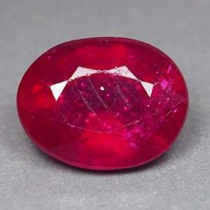 May include: An oval-cut gemstone with a deep, vibrant red color. The stone features a faceted surface that reflects light, creating bright white highlights across the top. The internal structure shows natural inclusions and a rich, saturated hue.