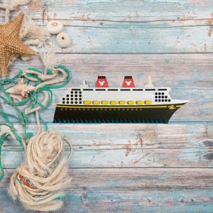 May include: A wooden toy cruise ship with red and yellow accents on a blue and white wood background. The ship has a Mickey Mouse head on the front.