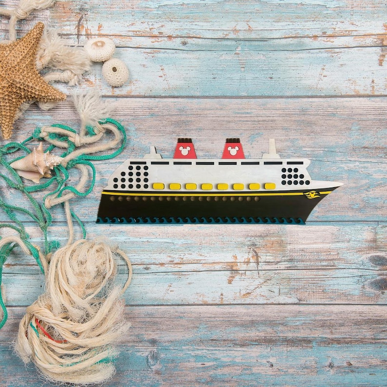 May include: A wooden toy cruise ship with red and yellow accents on a blue and white wood background. The ship has a Mickey Mouse head on the front.