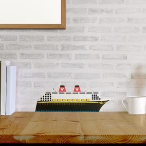 May include: A wooden model of a white cruise ship with red Mickey Mouse ears on the top. The ship has yellow portholes and a black hull. The model is sitting on a wooden table in front of a white brick wall.
