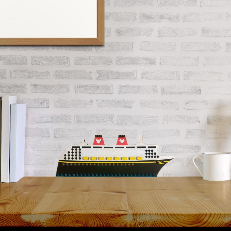 May include: A wooden model of a white cruise ship with red Mickey Mouse ears on the top. The ship has yellow portholes and a black hull. The model is sitting on a wooden table in front of a white brick wall.