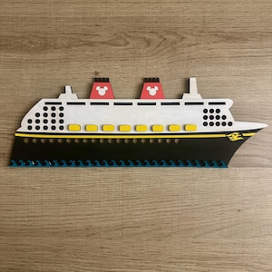 May include: A white and black cruise ship with yellow accents and red Mickey Mouse ears on the top. The ship is on a wood grain background.