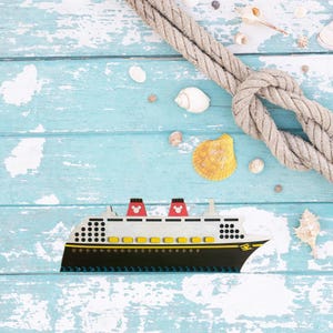May include: A toy cruise ship with red Mickey Mouse ears on the top of the ship. The ship is on a blue wooden background with a rope and seashells.