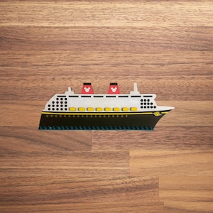 May include: A white and black cruise ship with red Mickey Mouse ears on the top of the ship. The ship is made of Lego blocks and is sitting on a wooden surface.