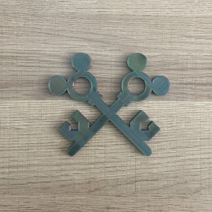 Könnte beinhalten: Zwei gekreuzte Schlüssel aus dunkelgrünem Kunststoff. Die Schlüssel haben oben ein kreisförmiges Design und unten ein detaillierteres Design.