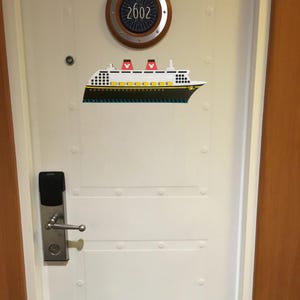 May include: White door with a silver door handle and a black digital lock. The door has a round porthole with the number 2602 on it. A white and black cruise ship sticker is on the door.