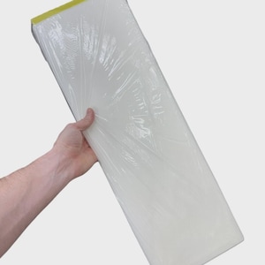 May include: A rectangular, white, flat object encased in clear, crinkled plastic wrap. The item features a distinct, solid yellow strip along one of its shorter edges. The overall shape is elongated and narrow, presenting a smooth, uniform surface.