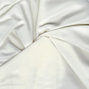 May include: A close-up of a white fabric with soft folds and wrinkles. The fabric is smooth and silky, with a subtle sheen.
