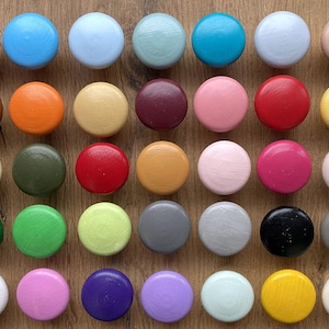 Könnte beinhalten: Sortiment von 36 runden Holzknöpfen in verschiedenen Farben, darunter Blau, Grün, Rot, Gelb, Braun, Rosa und Grau. Die Knöpfe sind in einem Gittermuster auf einer Holzoberfläche angeordnet.