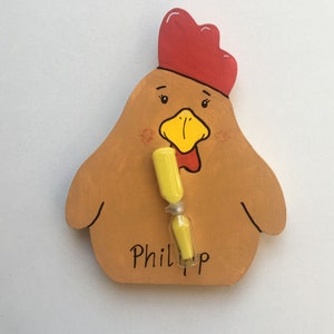 Könnte beinhalten: Ein hölzerner Hühner-Timer mit rotem Kamm, gelbem Schnabel und gelber Sanduhr. Der Name "Phillip" steht darunter. Das Huhn ist in Braun- und Beigetönen bemalt.