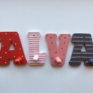 Könnte beinhalten: Holzbuchstaben, die den Namen "ALVA" buchstabieren, in verschiedenen Mustern bemalt. Die "A" sind rot mit weißen Punkten und grau mit weißen Streifen. Das "L" ist rosa mit roten und weißen Streifen. Das "V" ist rosa mit weißen Punkten. Jeder Buchstabe hat einen Holzgriff zum Aufhängen.