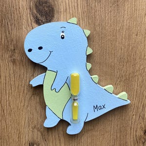 Könnte beinhalten: Ein blauer und grüner Holz-Dinosaurier mit einer gelben Sanduhr, die an seinem Bauch befestigt ist. Der Dinosaurier hat ein lächelndes Gesicht und der Name "Max" steht auf seiner Seite.