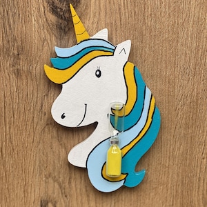 Könnte beinhalten: Ein weißes Holz-Einhorn mit blauem und gelbem Mähne und Schweif, das eine kleine gelbe Sanduhr hält.