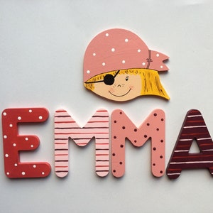 Könnte beinhalten: Holzbuchstaben, die den Namen "EMMA" in Rosa, Rot und Weiß mit Punkten und Streifen bilden. Über den Buchstaben befindet sich ein bemalter Holzkopf eines Mädchens mit einem rosa-weißen Tupfenbandana und einer Augenklappe.