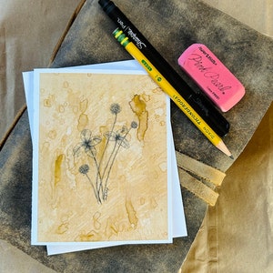 May include: A close-up of a watercolor painting of a clover plant on a light brown background. The painting is on a white card and is surrounded by a brown leather journal, a yellow pencil, and a pink eraser.
