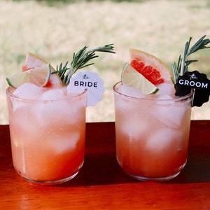 May include: Two pink cocktails with ice, grapefruit, lime, and rosemary garnishes. The drinks are labeled "The Bride" and "The Groom" with white and black paper flags.