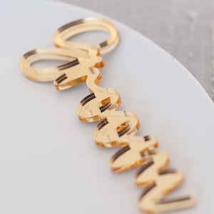 May include: A gold-colored acrylic name tag featuring the name "Grace" written in a cursive, flowing script font. The material has a reflective, mirror-like finish with visible layered edges, creating a three-dimensional appearance on the surface.