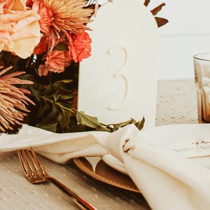 May include: A white ceramic table number with the number "3" in a modern font. The table number is surrounded by a floral arrangement with pink, orange, and red flowers. A gold fork and a white napkin are also visible.