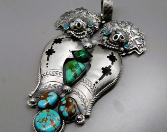 May include: A large, ornate silver pendant with turquoise and green stone accents. The pendant features a detailed floral design at the top and a stylized shape with cut-out details. The turquoise stones are set in silver bezels, and the overall design is intricate and eye-catching.