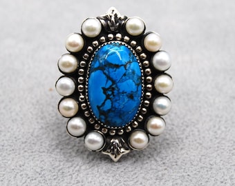 May include: Oval turquoise ring with a silver setting, surrounded by small white pearls. The turquoise stone is a vibrant blue with dark veining. The ring has a detailed, ornate design.