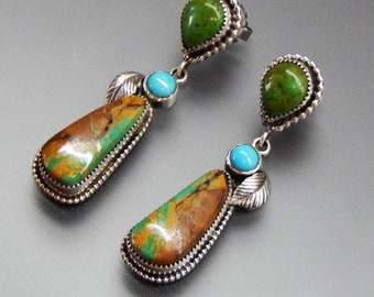 May include: A pair of silver earrings with teardrop-shaped stones. The earrings feature a combination of brown, green, and turquoise stones, with a silver leaf accent. The earrings have a detailed, textured silver border.
