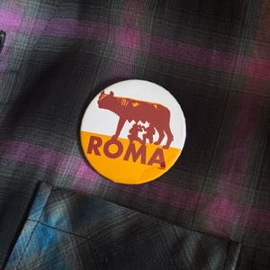 May include: A round button with a white and yellow background. The button features a brown illustration of a wolf nursing two babies, with the word "ROMA" in bold red letters. The button is set against a plaid fabric.