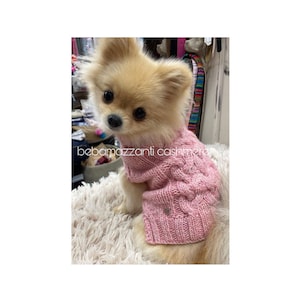 以下が含まれることがあります： 小さなふわふわの薄い茶色の犬が、ケーブル編みの模様の入ったピンクのニットセーターを着ています。セーターには、小さな銀色のハート型のボタンが前面についています。犬は白いふわふわの表面に座っています。