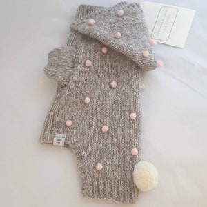 Op de afbeelding: Een grijze gebreide hoodie met roze pompoentjes en een witte pompoen op de staart. De trui heeft een label met de tekst "Handgemaakt met liefde".