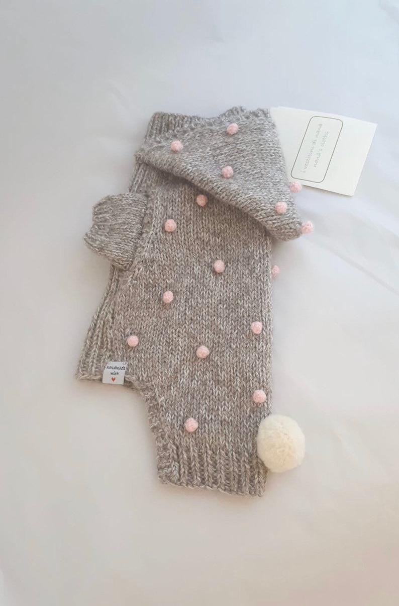 Op de afbeelding: Een grijze gebreide hoodie met roze pompoentjes en een witte pompoen op de staart. De trui heeft een label met de tekst "Handgemaakt met liefde".