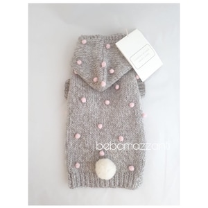 Op de afbeelding: Grijze gebreide hondentrui met een roze stippenpatroon en een witte pomponstaart. De trui heeft een capuchon en het label zegt "Handmade with love".