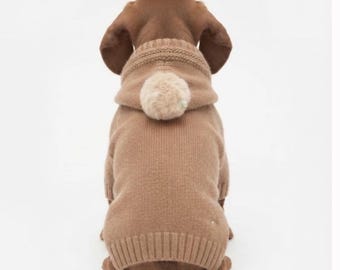 May include: A light brown knit dog sweater featuring a hood with a fluffy, round, cream-colored pom-pom attached to the back. The garment has a ribbed hem and ribbed cuffs, covering the back of a small dog with floppy ears and a short tail.