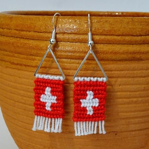 Puede incluir: Pendientes hechos a mano con un diseño tejido en rojo y blanco. Cada pendiente tiene una base rectangular roja con una cruz blanca, un borde blanco y flecos blancos. Los pendientes cuelgan de ganchos triangulares plateados.