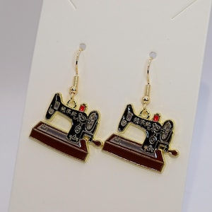 Peut inclure: Une paire de boucles d'oreilles pendantes dorées avec un motif de machine à coudre. Les boucles d'oreilles ont une machine à coudre noire et marron avec des accents dorés et des détails rouges. Sur fond blanc.