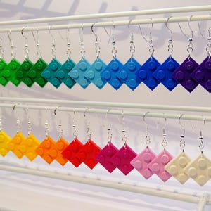 Könnte beinhalten: Eine Präsentation von 18 Paar Ohrringen, jedes mit einem farbenfrohen LEGO-Stein-Design. Die Ohrringe sind in einem Regenbogenmuster angeordnet, wobei jedes Paar eine andere Farbe aufweist. Die Ohrringe sind aus Silber und hängen an einem weißen Präsentationsständer.