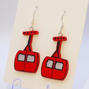 May include: A pair of red wooden earrings shaped like cable cars, with silver-colored ear wires. The cable cars have black outlines and two silver-colored windows. The earrings are displayed on a cream-colored background.