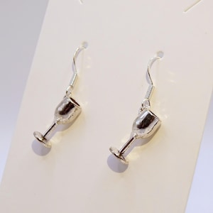 Può includere: Un paio di orecchini color argento a forma di calici da vino. Ogni orecchino ha un gancio per orecchie forate e un piccolo ciondolo a forma di calice dettagliato. Gli orecchini sono esposti su sfondo bianco.