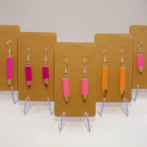 Peut inclure: Six paires de boucles d'oreilles en forme de crayon présentées sur du carton brun. Les boucles d'oreilles sont en argent avec un motif de crayon rose et orange.