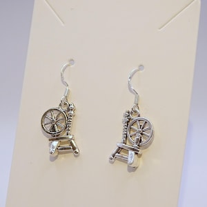May include: A pair of silver-colored earrings featuring a spinning wheel design. Each earring has a small wheel attached to a detailed frame, suspended from a curved ear wire. The earrings are displayed against a neutral background.