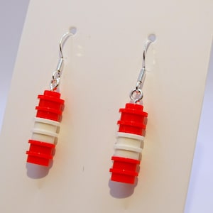 May include: A pair of dangle earrings featuring a stacked design of red and white cylindrical elements. The earrings are suspended from silver-colored fish hook ear wires. The earrings are displayed against a white background.