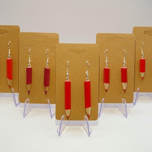 Op de afbeelding: Zes paar rode potloodvormige oorbellen getoond op kartonnen oorbellenstandaards. De oorbellen zijn zilver met een rode potloodhanger.