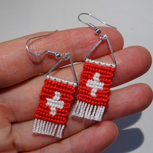 May include: A pair of handmade earrings featuring a red and white design, resembling the Swiss flag. The earrings have a rectangular shape with a white cross on a red background, and a fringe at the bottom. They are suspended from silver-colored hooks.