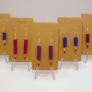 Op de afbeelding: Zes paar oorbellen in de vorm van een potlood, gepresenteerd op bruin karton. De oorbellen zijn in drie kleuren: paars, blauw en rood.