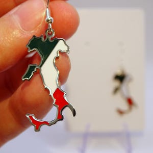 Peut inclure: Boucles d'oreilles argentées en forme de la péninsule italienne, avec de l'émail vert, blanc et rouge aux couleurs du drapeau italien. Les boucles d'oreilles sont suspendues à des crochets argentés. Une autre boucle d'oreille est visible en arrière-plan.
