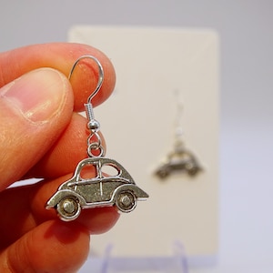 May include: Silver-toned car-shaped earrings. The earrings feature a detailed design of a classic car with wheels, windows, and a curved roof. The earrings are suspended from a silver hook.