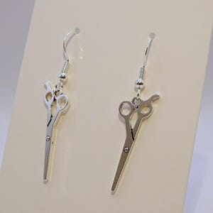 Op de afbeelding: Een paar zilveren oorhangers met een schaar-bedel. De oorbellen zijn op een witte achtergrond.